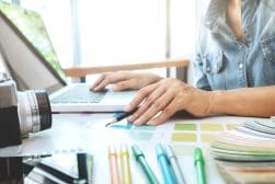 A person using a laptop is holding a pencil, surrounded by color swatches and various colored pens. A camera is placed next to them on the desk.
