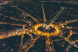 An aerial nighttime view of the Arc de Triomphe in Paris, surrounded by illuminated streets and city lights.