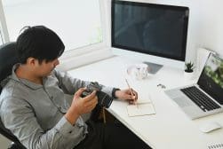A man is sitting at a desk holding a camera and writing in a notebook. The desk has a monitor, laptop, headphones, and a small plant.