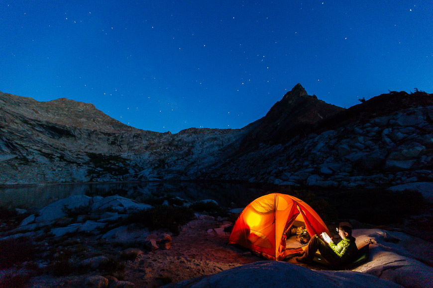 A camper reading inside an illuminated tent by a mountain lake under a starry night sky.