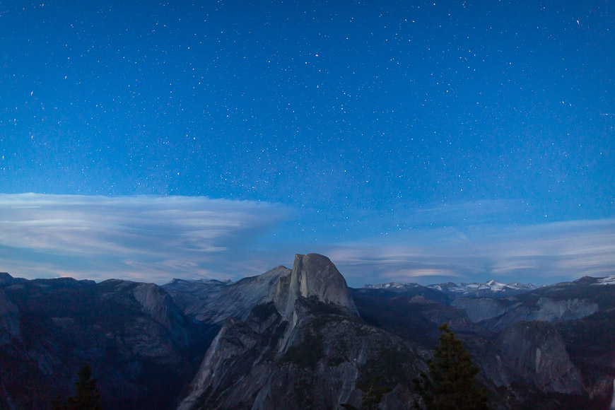 A nighttime view of Half Dome under a starry sky in Yosemite National Park, with clouds and mountain ridges visible in the distance.