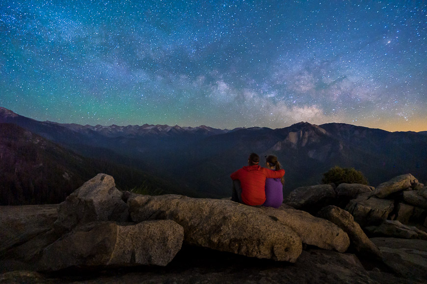 Two people sit on large rocks, gazing at a star-filled night sky over a mountainous landscape.
