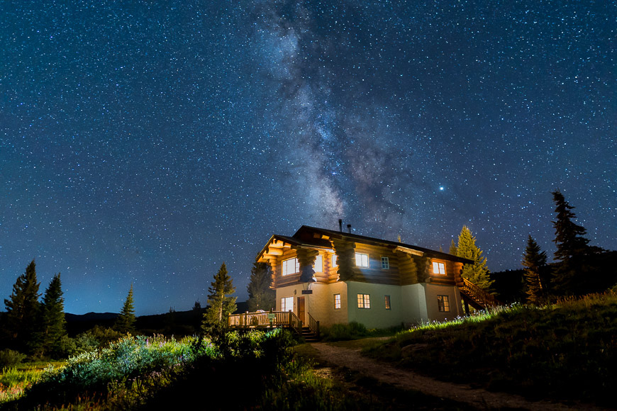 Log cabin with lights on, set in a lush meadow under a starry night sky with the Milky Way visible above.