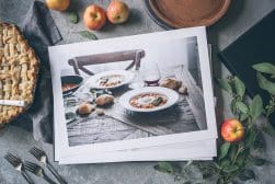 A tabletop with an open book showing a photo of a meal, surrounded by apples, a pie, utensils, and leafy branches.