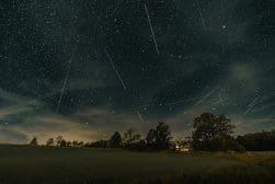 A night sky filled with numerous shooting stars over a landscape of trees and a house with lights on.