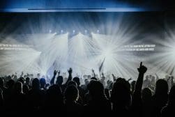 A crowd at a concert with raised hands silhouetted against bright stage lights and beams in the background.