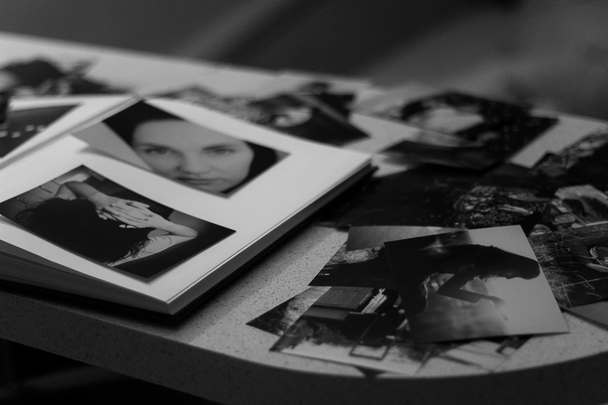 Black and white photograph of an open photo album on a table surrounded by scattered photos. The album contains pictures of a woman.