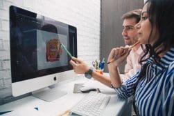 Two people sitting at a desk, reviewing a graphic design on a desktop computer; one is holding a green pencil pointing at the screen, and the other is holding an orange pencil near their face.