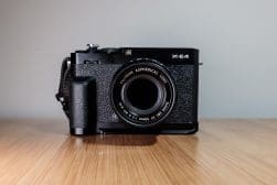 A black Fujifilm X-E4 camera with a Fujinon aspherical lens placed on a wooden surface against a plain background.