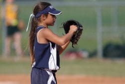 A young girl in a navy blue softball uniform and visor holds a softball glove, preparing to catch a ball on a field.