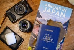 A camera, a camera lens, a book titled "Abroad in Japan," an Australian passport, wallets, and AirPods on a wooden surface.