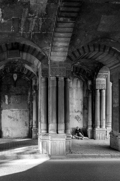 A person sits on the ground under a stone archway in a dimly lit area with decorative columns.