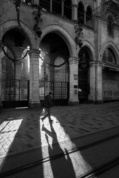 A person walks on a sunlit street casting a long shadow, with an ornate building featuring arches in the background.