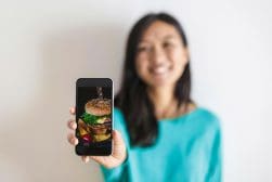 A person in a turquoise shirt is smiling and holding a smartphone displaying a close-up image of a burger.