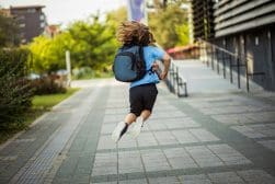 A child with a backpack is running and mid-air on a paved pathway next to a building, surrounded by greenery.