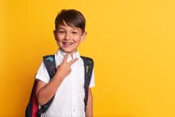 A young boy wearing a white shirt and backpack smiles, holding up a peace sign with his fingers against a solid yellow background.