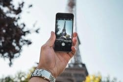 A person holds a smartphone in front of the Eiffel Tower, capturing the tower's image on the screen.