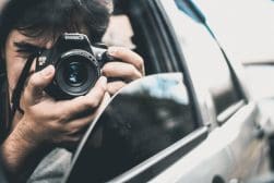 A person leans out of a car window, holding and aiming a Canon camera directly at the viewer.