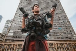A person stands in front of a tall building, holding multiple cameras and lenses, with more cameras hanging around their neck.