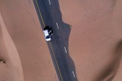 Aerial view of a white car driving on a paved road through sandy desert terrain.