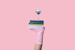 A hand in a pink sock holds a stack of books with a cocktail glass on top, against a pink background.