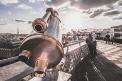A telescope mounted on a city observation deck overlooks an urban landscape with the Eiffel Tower visible in the distance under a partly cloudy sky.