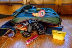 A green bag spills open on a wooden floor, revealing a camera with a lens, a wristwatch, a Swiss Army knife, a small yellow pouch, a wallet, and camera batteries.