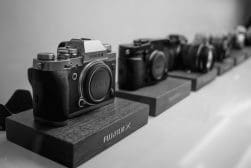 A row of Fujifilm cameras is displayed on a shelf, with the closest camera prominently featured in the foreground.