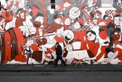 A person walks a dog along a sidewalk in front of a large, colorful mural featuring numerous stylized characters and various activities in shades of red, white, and black.
