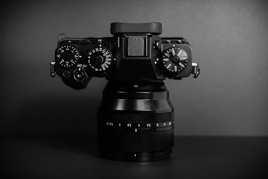 Black and white photo of a digital camera with dials on top and a zoom lens, positioned against a gray background.