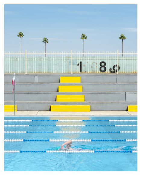 A person swims in a blue pool with lane dividers. Yellow and gray bleachers and a fence are in the background, with numbers "1 8" visible. Palm trees and a clear sky are above the fence.