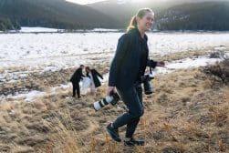 A photographer, holding two cameras, walks up a grassy slope with a snow-covered field and mountains in the background. In the distance, a couple stands together.