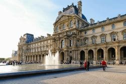 People gather around a large fountain in the courtyard of the Louvre Museum, a grand historic building with ornate architecture, under a clear blue sky.