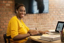 Person smiling, wearing earphones, and working on a laptop at a table with bricks wall in the background. Various papers are on the table.