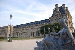 A statue of a woman seated in front of the Louvre Museum in Paris, France, with the museum building visible in the background.