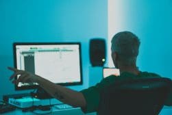 Person with short hair sits in front of a computer with a large screen, surrounded by audio equipment, pointing towards the screen. The room is bathed in blue light.