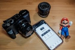 A camera, two lenses, a smartphone displaying a camera app, and a small Mario figurine are arranged on a wooden surface.