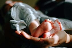 Close-up of an adult hand cradling the feet of a baby, who is lying down and wrapped in a blanket.