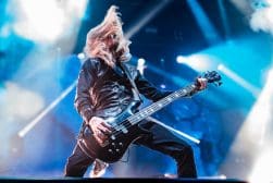 A musician with long hair plays an electric guitar on stage under vibrant blue and white lights.