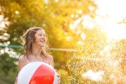A young girl with wet hair holds a beach ball and smiles in sunlight, with water droplets visible around her.