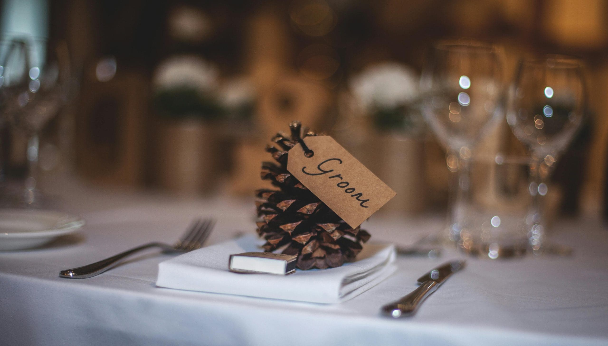 A pinecone with a "Groom" label serves as a table place card, placed on a white napkin next to utensils and glassware on a set dining table.