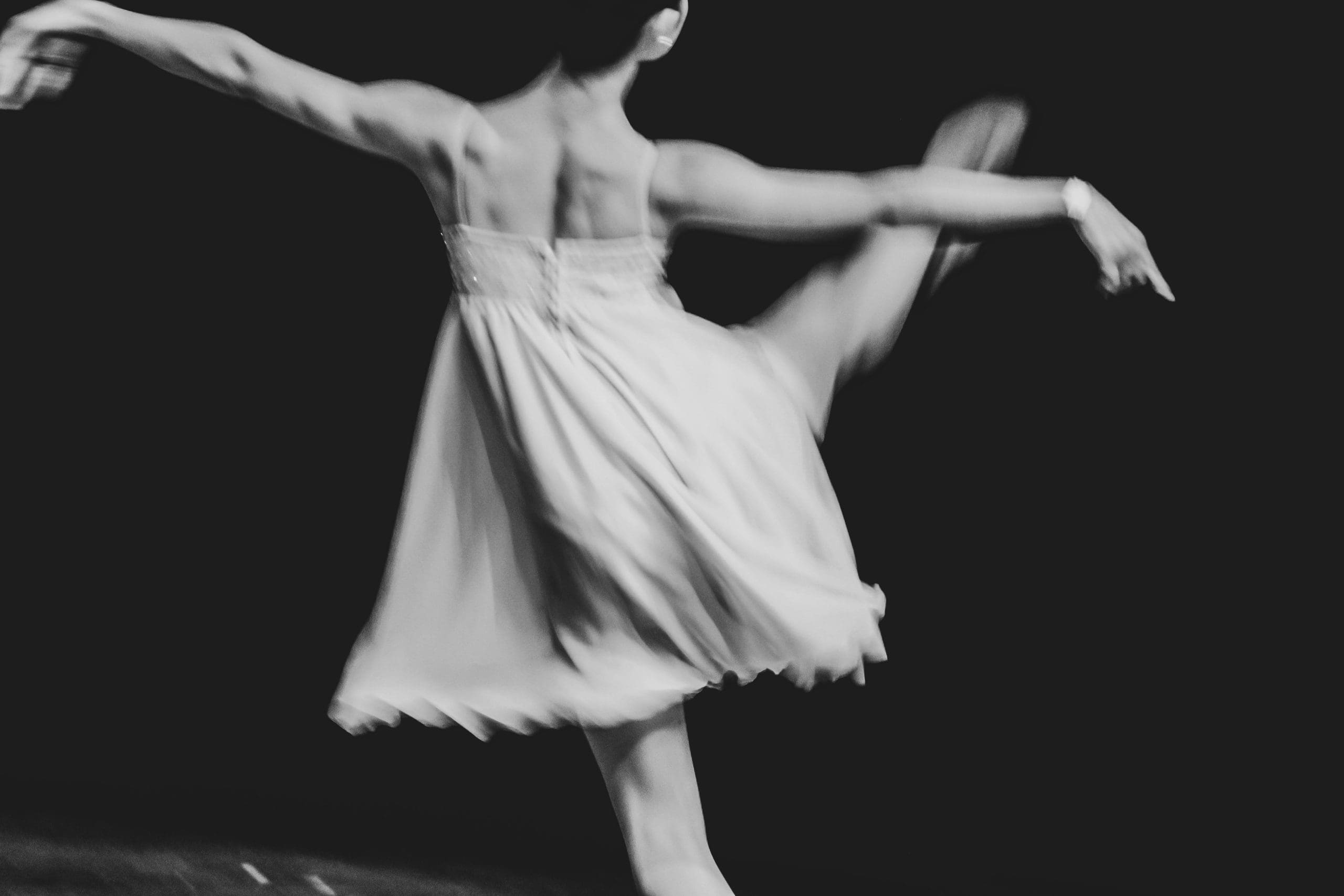 A female dancer in a flowing dress balances on one leg with the other raised and arms extended in a graceful pose against a black background.