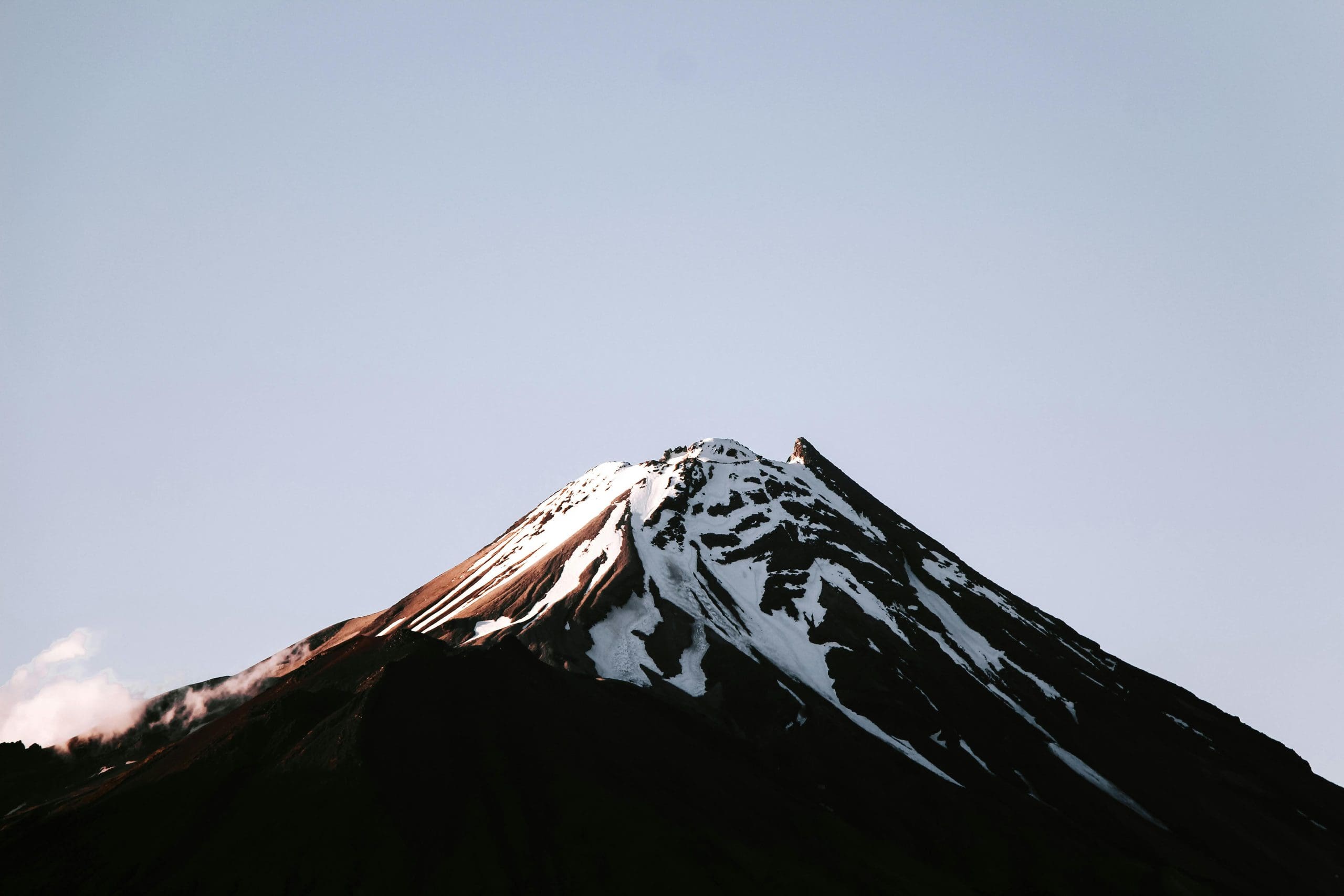Snow-topped mountain peak with a clear sky background. Sparse snow patches adorn the rugged, dark rock surface.