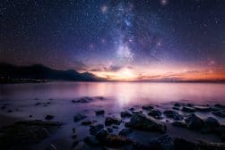 A calm, rocky shoreline under a starry night sky, with the Milky Way galaxy visible, and mountains in the background at twilight.