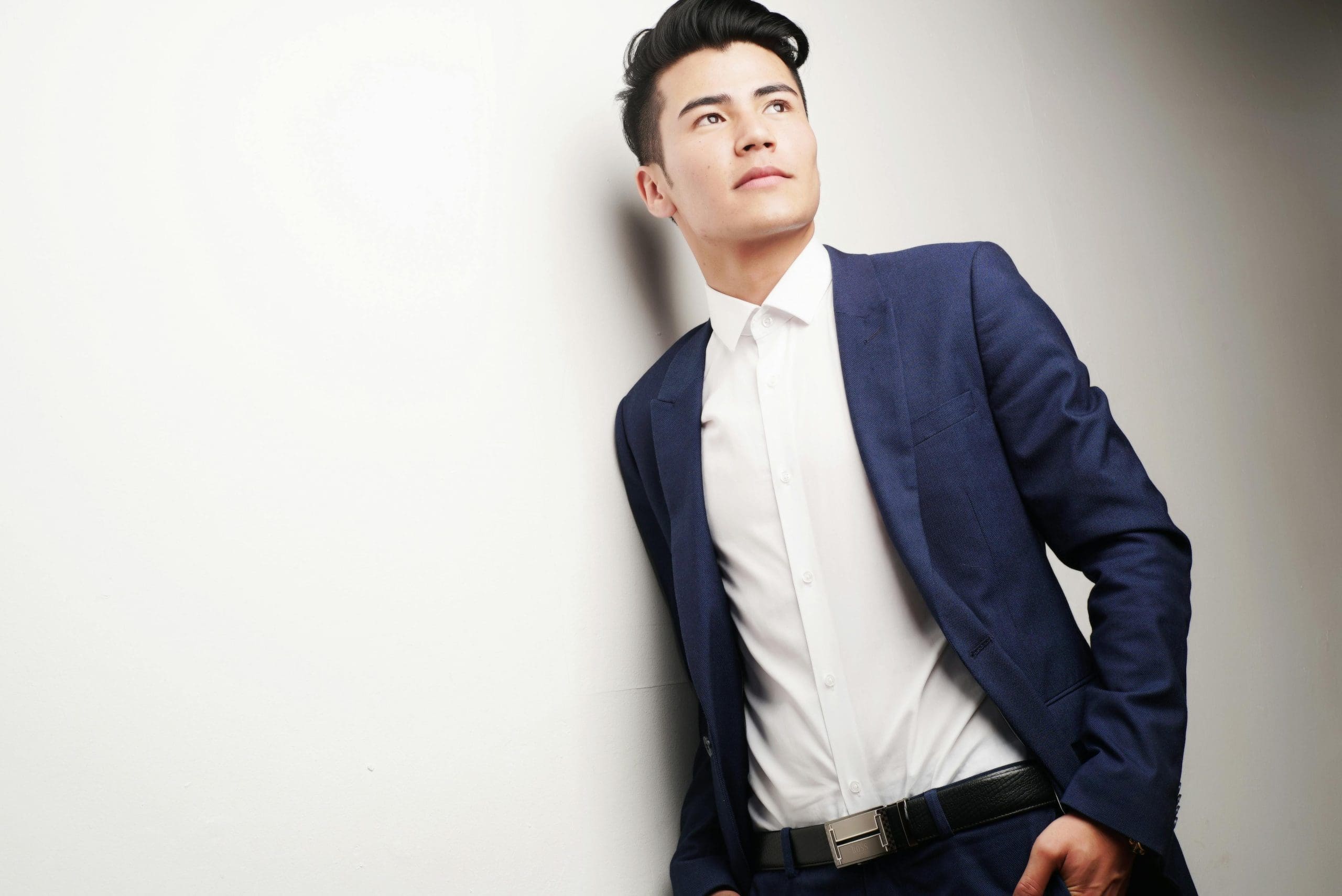A man in a blue suit and white dress shirt leans against a white wall, looking upwards with his hand in his pocket.