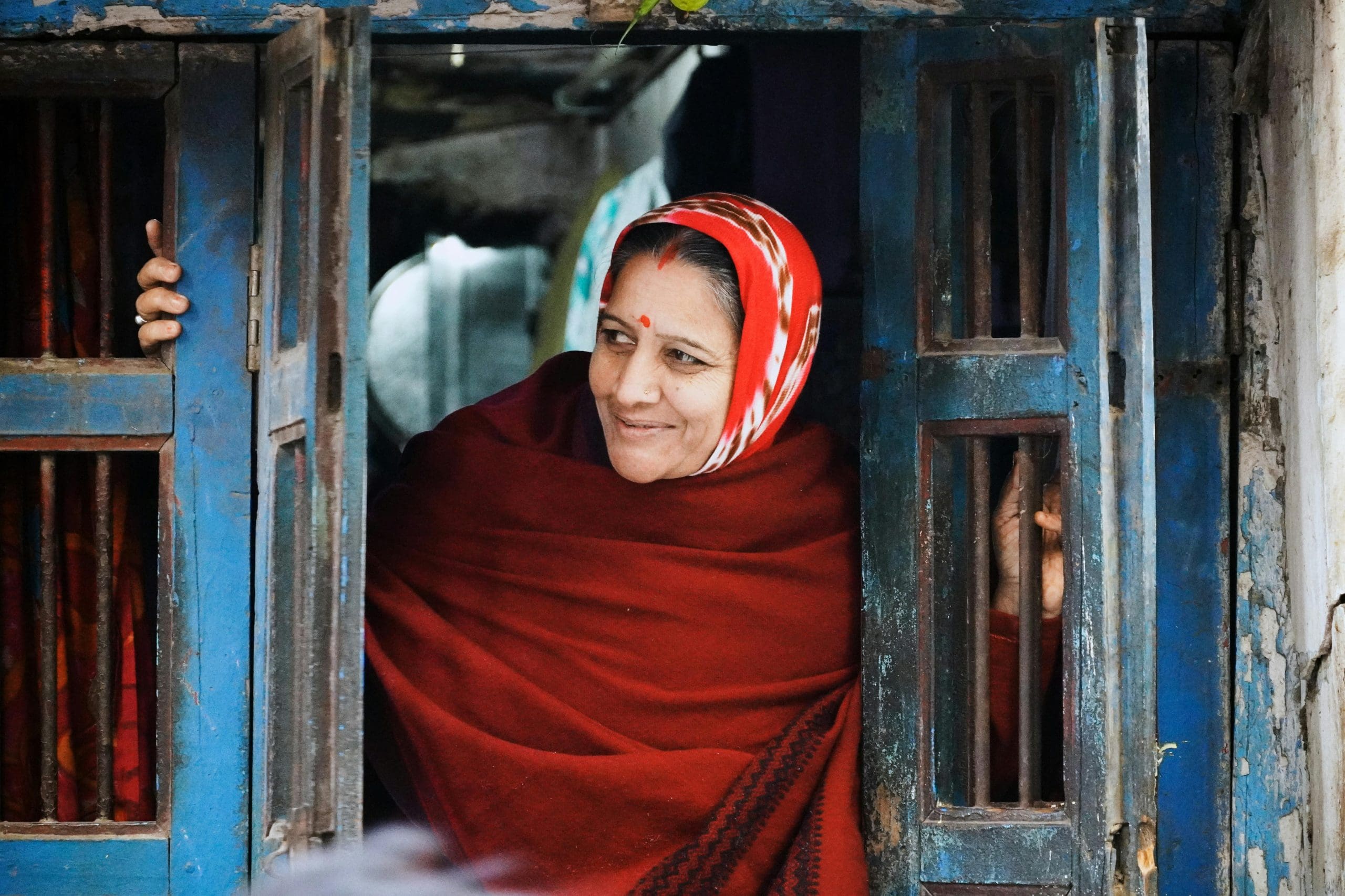 A woman wearing a red shawl and headscarf stands at an open window, holding the blue wooden shutters.