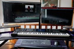 A dual monitor setup with video editing software on the larger screen and music production software on the smaller screen. Below, a MIDI keyboard, computer keyboard, and mouse sit on the desk.