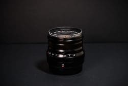 A Fujifilm camera lens with a lens protector, positioned upright against a dark background. The aperture ring indicates various f-stops.