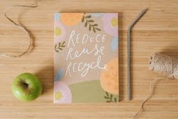 A notebook with the text "Reduce Reuse Recycle" on a floral cover, next to a green apple, metal straw, twine, and string on a wooden surface.