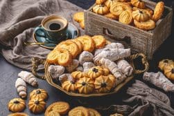 A variety of cookies displayed on a tray and in a wooden box, accompanied by a cup of coffee on a saucer with a spoon, surrounded by a rustic cloth.